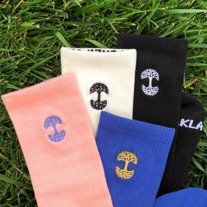 A collection of Oaklandish Embroidered Logo Socks is scattered on a green grass background. The socks are in pink, white, black, and blue, each featuring a unique embroidered symbol resembling either a tree or mushroom in various colors. The word "KLA" is partly visible on the edge of one of the black crew socks.