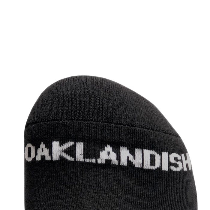Close-up of the toe section of a black crew sock from the Oaklandish brand's Embroidered Logo Socks collection. The sock features "OAKLANDISH" in white, capital letters, prominently displayed near the front. The background is plain and light-colored, offering high contrast to the black sock and white text. These socks are available in adult sizes.
