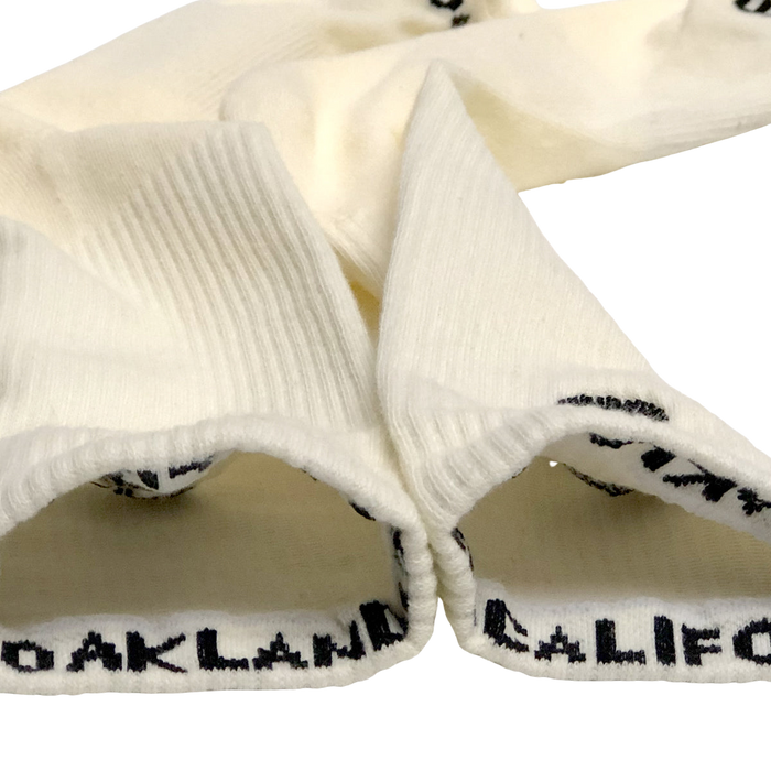 Two off-white cotton crew socks from Oaklandish, known as Embroidered Logo Socks, are laid out with their cuffs turned inside out. The cuffs have a ribbed texture and feature the text "OAKLAND" in black capital letters at the interior edge. These socks are positioned so that the text is partially visible.