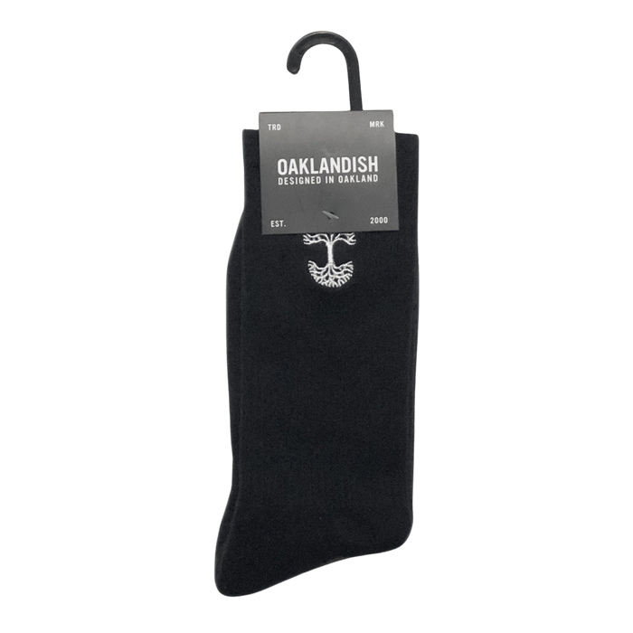 A single black Embroidered Logo Sock from Oaklandish is displayed on a hanger, with a label attached at the top. The label reads "OAKLANDISH DESIGNED IN OAKLAND" along with "TRD MRK" on either side. The year "EST. 2000" is also mentioned. An emblem resembling a tree appears on the adult-sized Oaklandish sock below the label.