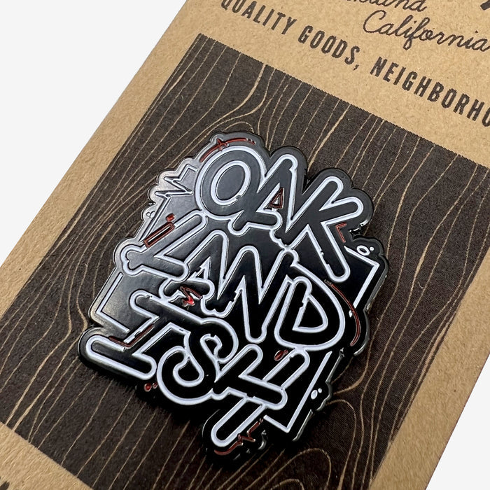 The Enamel Pin - Movement by Oaklandish features the stylized text "OAKLANDISH" in white letters on a black enamel background. Displayed on a cardboard backing that imitates a wood grain texture, it also includes partial text reading "QUALITY GOODS, NEIGHBORH...". This pin is perfect for accessorizing any outfit.