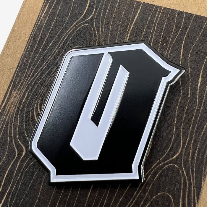 Close-up image of the Oaklandish Enamel Pin - Official, featuring a metallic emblem shaped like the letter "O" with a sleek black and silver design. The enamel lapel pin is placed on a surface with a dark wood grain pattern, highlighting the contrast between the accessory's colors and the textured background.