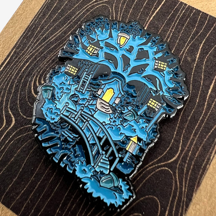 An intricate Enamel Pin - Treehouse by Oaklandish depicts a serene night scene with a treehouse. The tree in this Oakland-inspired design is adorned with lantern-like windows, and below it, a wooden bridge spans across a small creek. This accessory features shades of blue, capturing a calm, whimsical atmosphere.