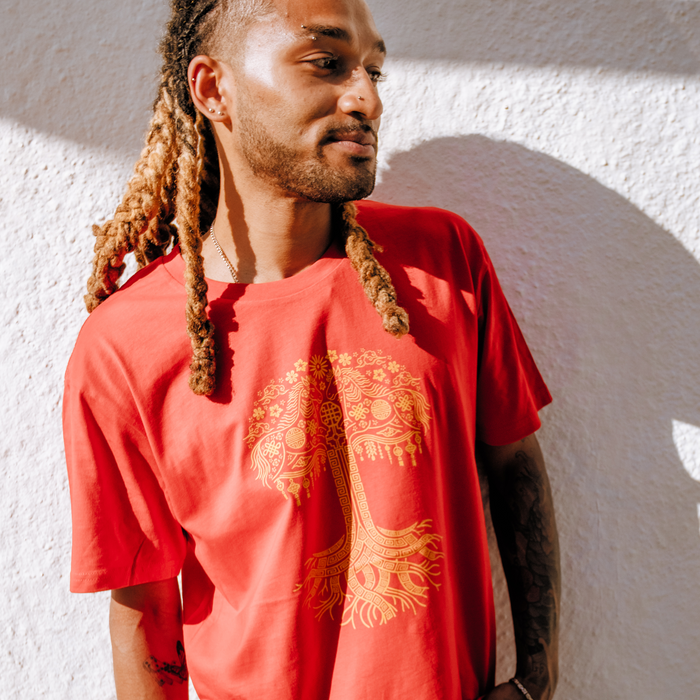 A person with long, textured hair and a beard stands against a sunlit white wall, wearing the Oaklandish Fire Horse Tee with a gold tree design. They smile softly to the side, showing a tattoo on their arm.