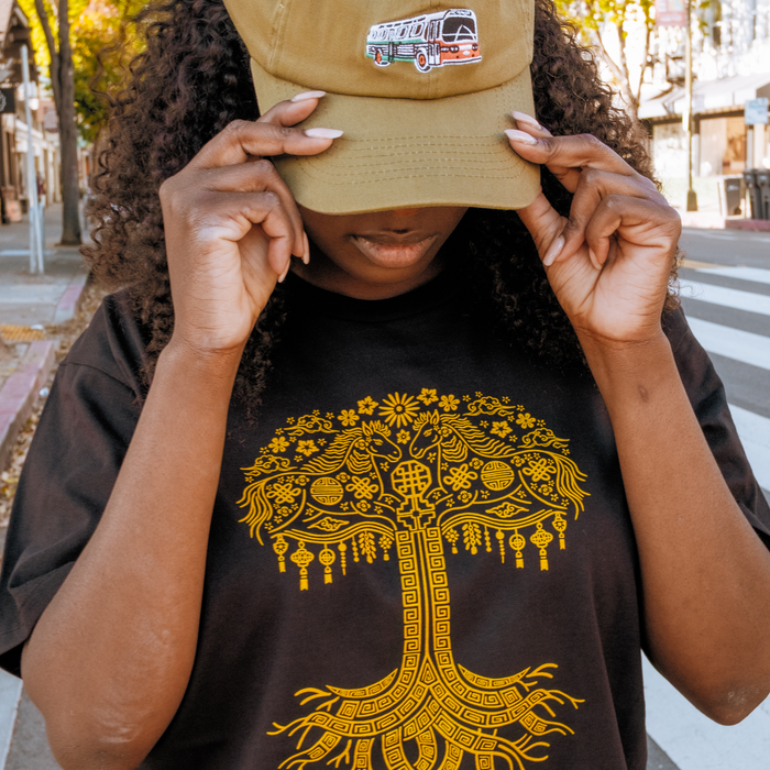 A person with curly hair holds a tan cap with a bus patch while wearing the Oaklandish Fire Horse Tee, a classic fit 100% cotton shirt featuring an intricate yellow tree design. The backdrop is a sunlit street lined with trees and buildings.