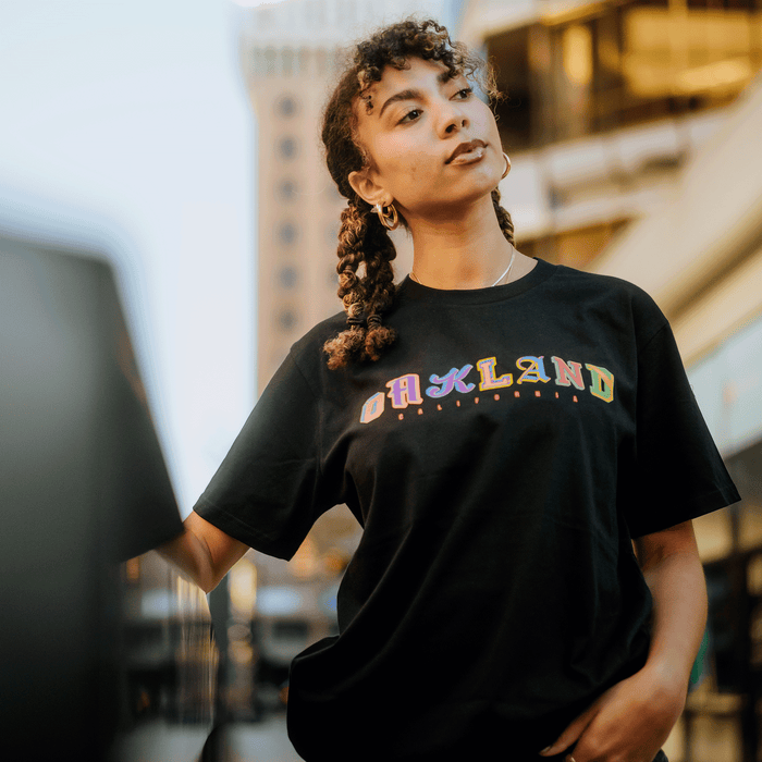 A woman with curly hair styled in braids and gold hoop earrings stands outdoors in a city, wearing Other’s First Team Tee—a black heavy-weight cotton T-shirt with “OAKLAND” in colorful letters. Blurred buildings and warm sunlight fill the background as she gazes thoughtfully to the side.