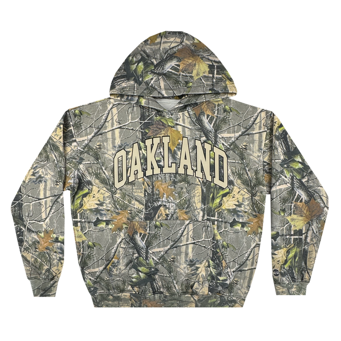The Oaklandish Foliage Hoodie features a green, brown, and gray wood camo pattern, bold cream "OAKLAND" lettering, an embroidered tree logo, front pocket, and adjustable drawstring hood.