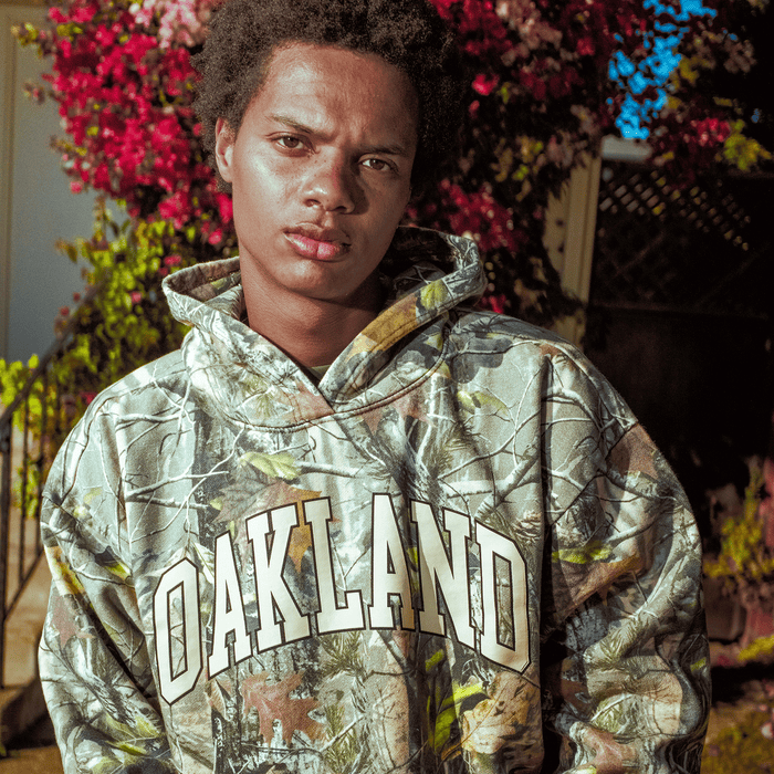 A young person stands outside in sunlight, wearing the Oaklandish Foliage Hoodie with "OAKLAND" in bold white letters and the Oaklandish tree logo. Surrounded by pink flowers and greenery, they have short curly hair and a serious expression.