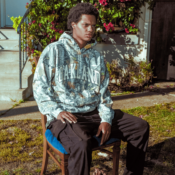 A person with short curly hair sits outdoors on a wooden chair with a blue cushion. They wear the Oaklandish Foliage Hoodie and black pants. Behind them are steps, green bushes, and vibrant pink flowers in sunlight.
