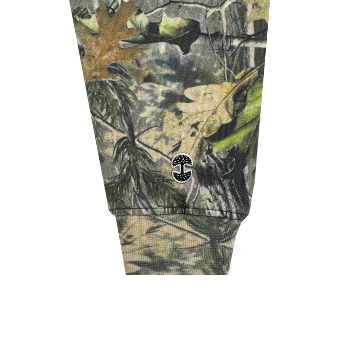 A close-up of the Foliage Hoodie by Oaklandish shows a sleeve cuff with a wood camo pattern in green, brown, and tan. Near the cuff is a small black embroidered Oaklandish tree logo with two mirrored 'C' shapes in a circular design.