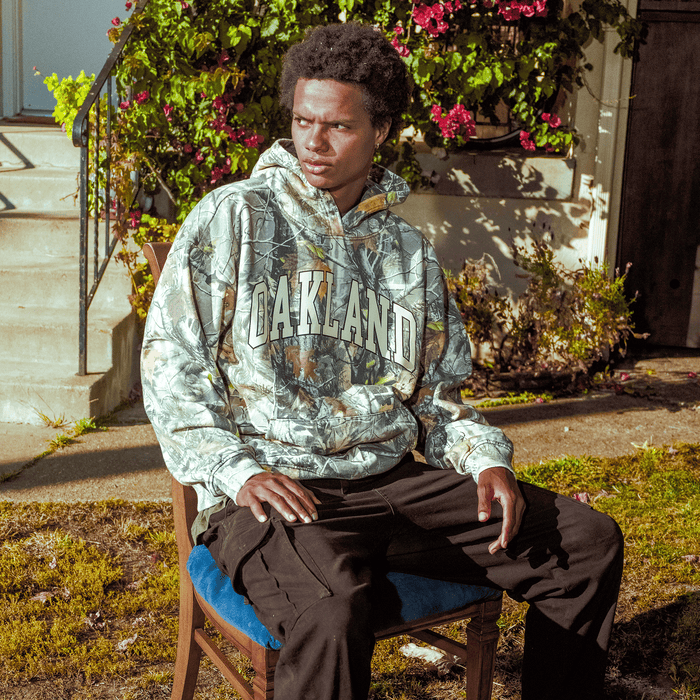 A young man sits outside on a wooden chair, wearing the Oaklandish Foliage Hoodie and black pants. Sunlight highlights his face as greenery and flowering bushes frame the concrete steps by the building entrance.