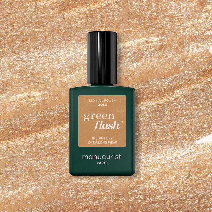 A bottle of Manucurist Gold gel polish rests on a sparkling gold glitter background, with the label showing "Gold" and Manucurist branding.
