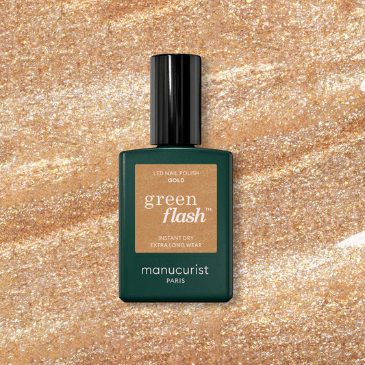A bottle of Manucurist Gold gel polish rests on a sparkling gold glitter background, with the label showing "Gold" and Manucurist branding.