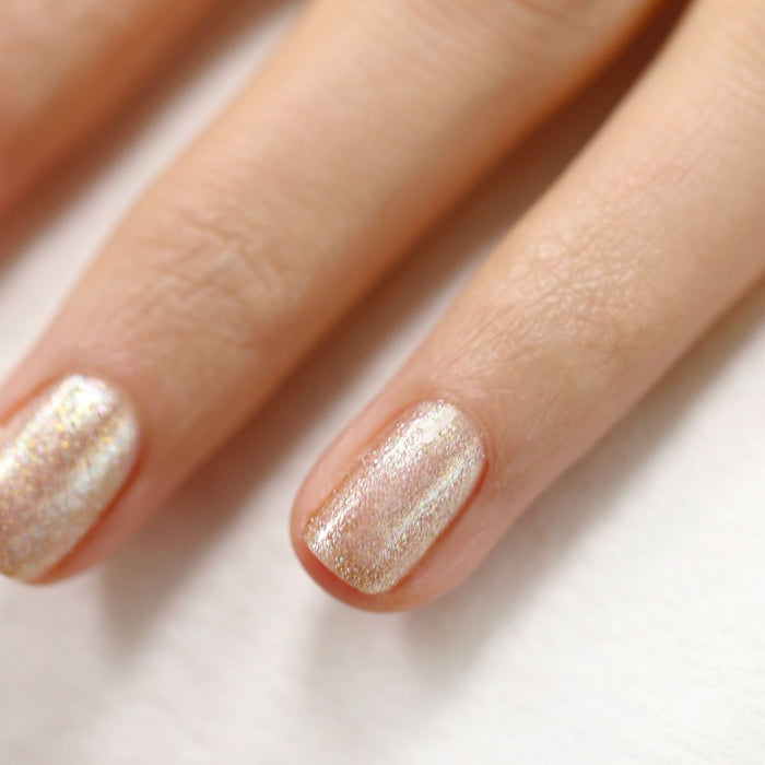 A hand with neatly manicured nails painted in Manucurist’s Gold shimmery, light pearlescent polish touched with gold glitter gel rests elegantly on a white surface.