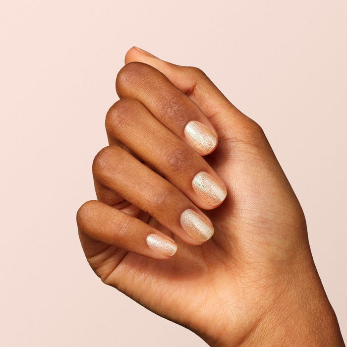 A hand with medium brown skin tone displays nails painted with Manucurist Gold, a shimmery light beige glitter polish, against a plain light background.