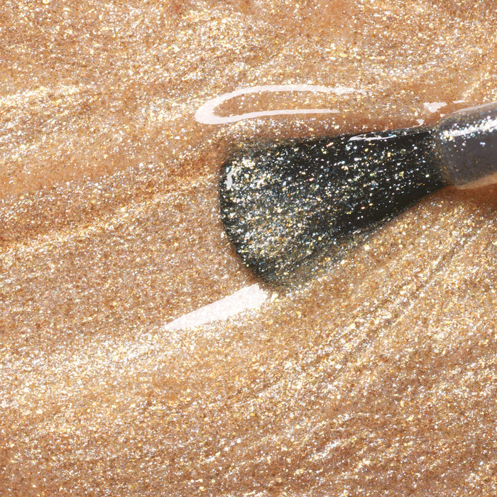 A close-up of a Manucurist Gold nail polish brush applying fine gold glitter gel polish over a glossy golden surface.