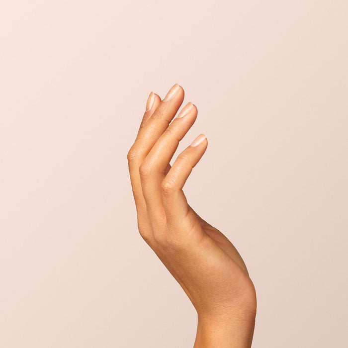A hand with neatly manicured nails—finished in Manucurist’s Gold gel polish—is raised against a plain, light beige background, fingers gently curved and palm facing inward.
