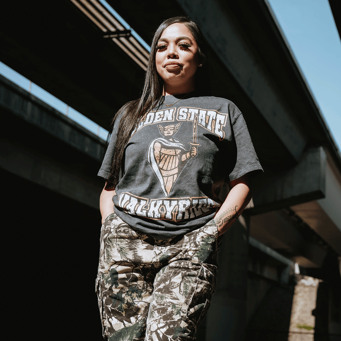 A woman with long dark hair stands confidently under a sunlit bridge, wearing the GSV Golden State Valkyries Guardian Stone Wash Heavy Tee and camouflage pants. Blue sky and shadows appear in the background.