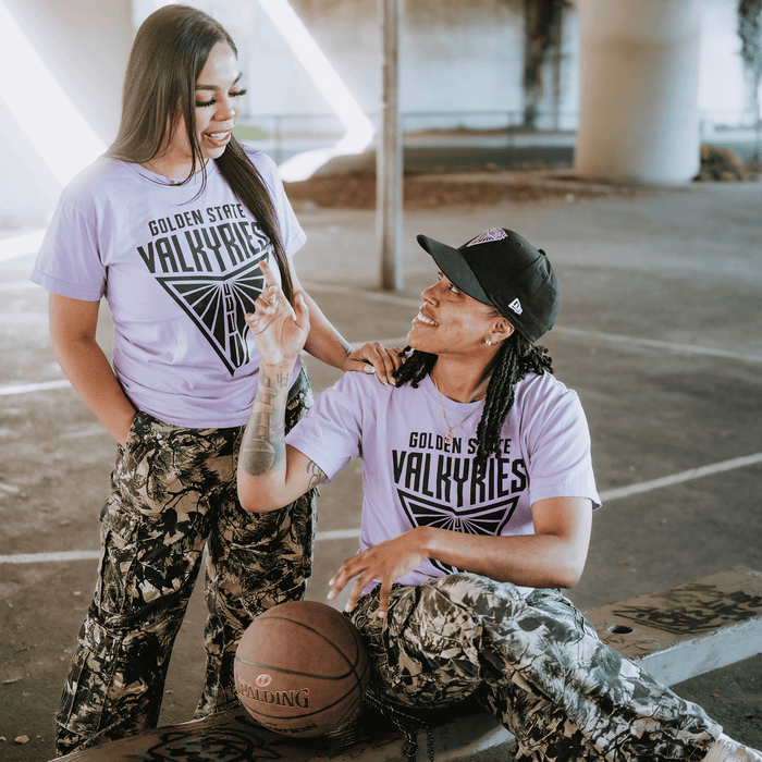 Two people stand on a basketball court under a bridge, both in matching GSV Golden State Valkyries Primary Premium Tees and camouflage pants. One sits with a basketball, smiling at the other, who stands nearby and smiles back.