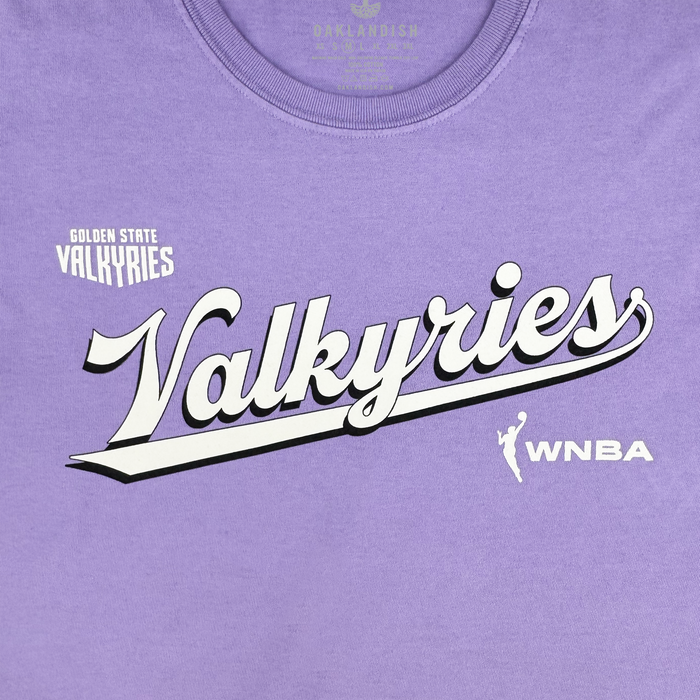 The GSV Golden State Valkyries Retro Script Premium Tee is a lavender organic cotton shirt featuring bold white "Valkyries" script with a black shadow, "Golden State Valkyries" above in small print, and the white WNBA player logo on the right.
