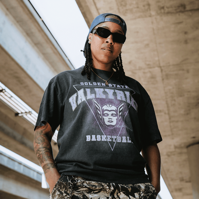 A person with long braids, sunglasses, and a cap stands under an overpass. They wear the GSV Golden State Valkyries Surge Stone Wash Heavy Tee and camouflage pants, exuding confidence with tattoos on one arm and hands in pockets.