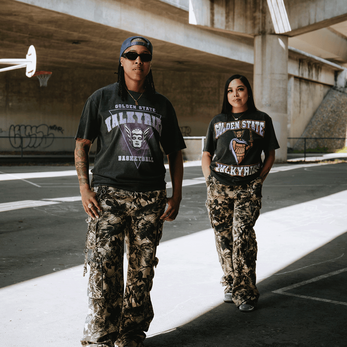 Two people stand confidently on a basketball court under an overpass, both wearing GSV Golden State Valkyries Surge Stone Wash Heavy Tees and camouflage pants, as sunlight casts sharp shadows on their standout style.