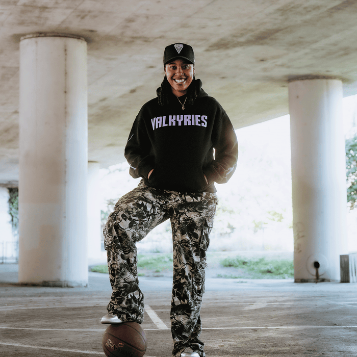 A person smiles under a bridge, wearing a black GSV Golden State Valkyries Violet Lightning Hoodie and camouflage pants with a black cap. They rest one foot on a basketball on concrete, with columns and greenery in the background.