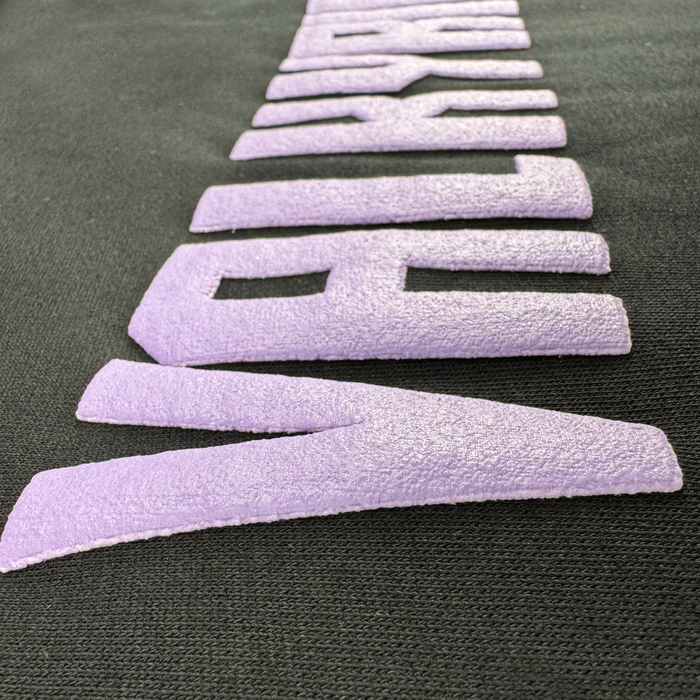 Close-up of textured, raised violet letters spelling "NEW WAVE" diagonally on a black mid-weight fleece fabric. The soft, suede-like text stands out against the smooth material of the GSV Golden State Valkyries Violet Lightning Hoodie.