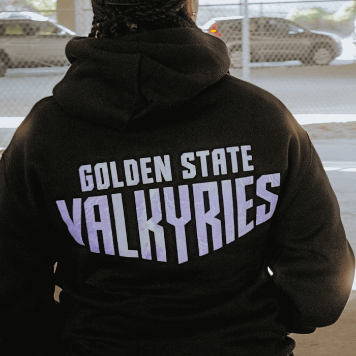 A person wears the GSV Golden State Valkyries Violet Lightning Hoodie, an officially licensed black mid-weight fleece pullover with "GOLDEN STATE VALKYRIES" in bold on the back, seen outdoors near a chain-link fence and blurred cars.