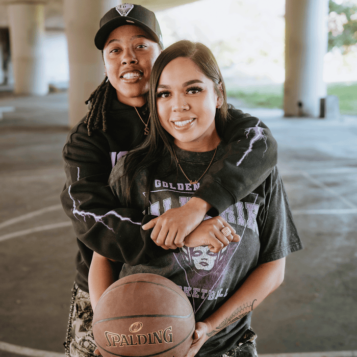 Two people smile on a basketball court under an overpass. One wears a GSV Golden State Valkyries Violet Lightning Hoodie, standing behind with arms around the other, who holds a Spalding basketball as daylight streams in.