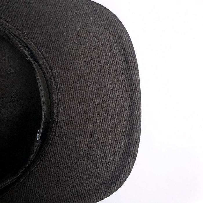 Close-up image of the underside of a Greenback Snapback's brim. The cap's fabric texture and stitched lines are clearly visible. The image focuses on the front part of the brim with a white background on the right side. This headwear style, from Oaklandish, combines both comfort and urban flair.