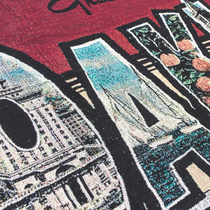 Close-up of the Greetings From Oakland Throw by Oaklandish, featuring a graphic design on a burgundy fabric with the word "SAN" in large, stylized letters. Each letter is filled with a vintage postcard-inspired collage of colorful images depicting scenes like architecture, landscapes, and flowers. The vibrant design has a textured appearance and uses recycled fibers.