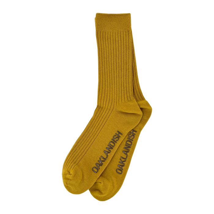 A pair of mustard yellow Grip Crew Socks by Oaklandish, featuring "OAKLANDISH" in dark gray capital letters along the sides. These yellow accessories have a ribbed texture from top to ankle, with reinforced toe and heel areas slightly darker than the rest. They are made from a cotton polyester spandex blend.