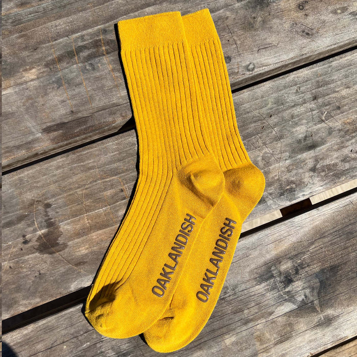 A pair of Oaklandish Grip Crew Socks in vibrant yellow with a ribbed texture are laid out on a weathered wooden surface. "OAKLANDISH" is printed in bold, black letters on the soles. These stylish yellow accessories, made from a blend of cotton, polyester, and spandex, are captured in natural sunlight.
