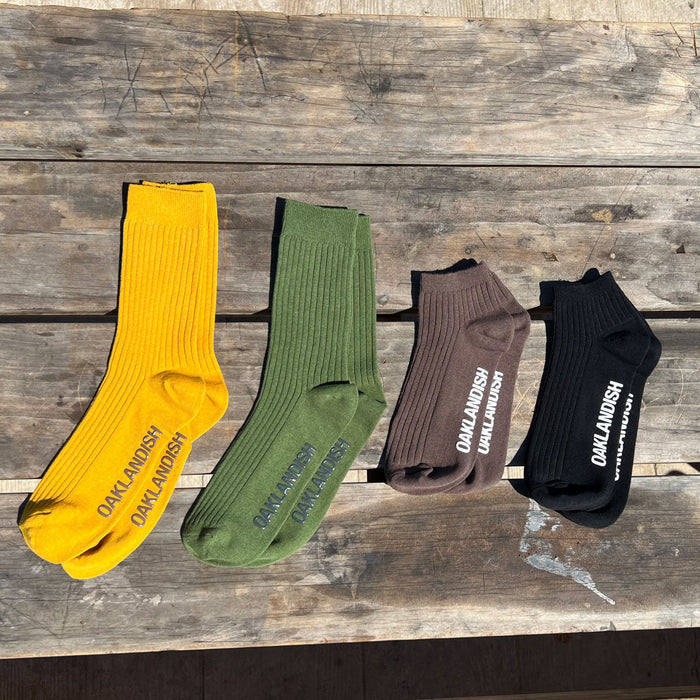 A set of four Grip Crew Socks by Oaklandish displayed on a rustic wooden surface. These ribbed cotton, polyester, and spandex socks come in yellow, green, brown, and black. Each pair prominently features the word "OAKLANDISH" printed on the side in contrasting colors. Sunlight highlights their texture against the wood.