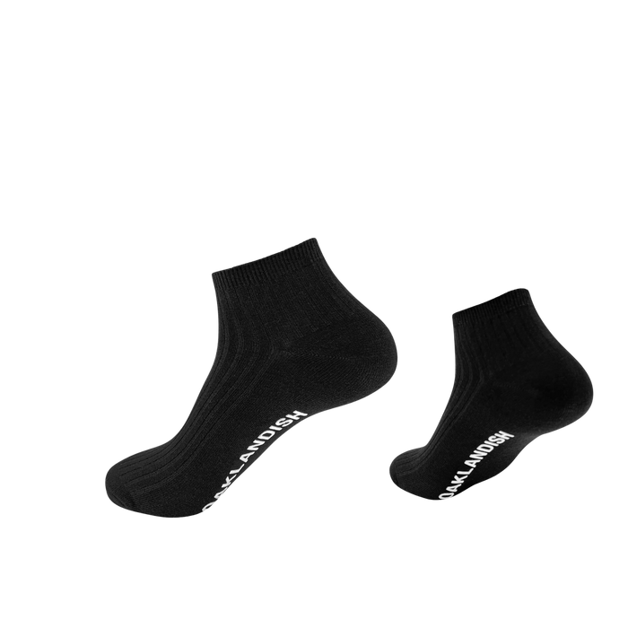 The Grip Low Crew Sock by Oaklandish is displayed against a simple white background. These black socks feature a ribbed texture and have "SOCKLANDISH" written in white text along the outside of the arch. The socks appear to be slightly floating, casting shadows on the surface below them.