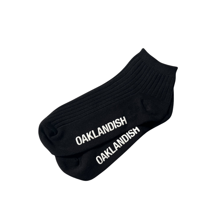 A pair of Grip Low Crew Socks from Oaklandish, featuring a black, ribbed design with "OAKLANDISH" printed in white capital letters along the top, are laid flat against a plain white background.