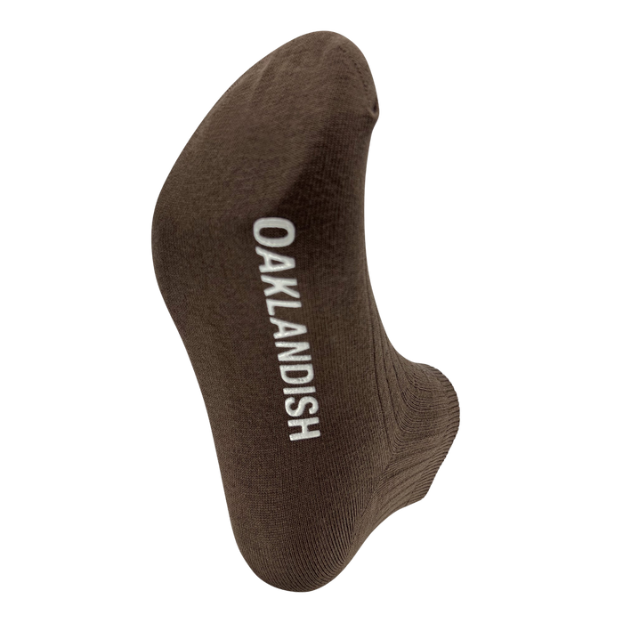The Oaklandish Grip Low Crew Sock is a single brown cotton low crew sock with the brand name "OAKLANDISH" in white capital letters running vertically. Photographed on a white background, the image highlights the heel and toe portions of the sock, emphasizing its fabric texture and simple design.
