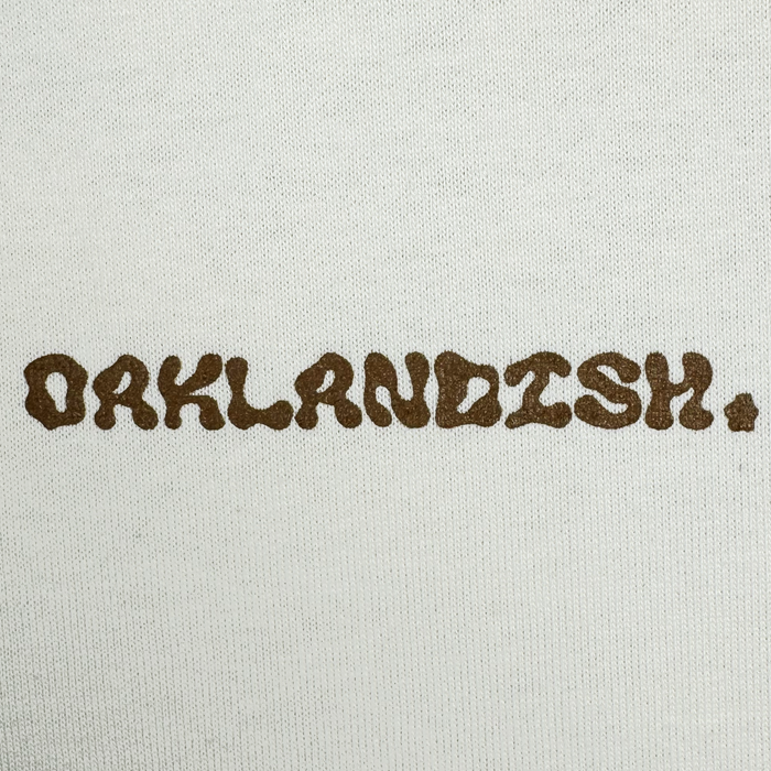 The image displays the word "OAKLANDISH" in textured brown letters on a plain white background, reminiscent of the Grow Your Roots Heavy Tee from Oaklandish, designed for a boxy fit that's popular in men's sizing.