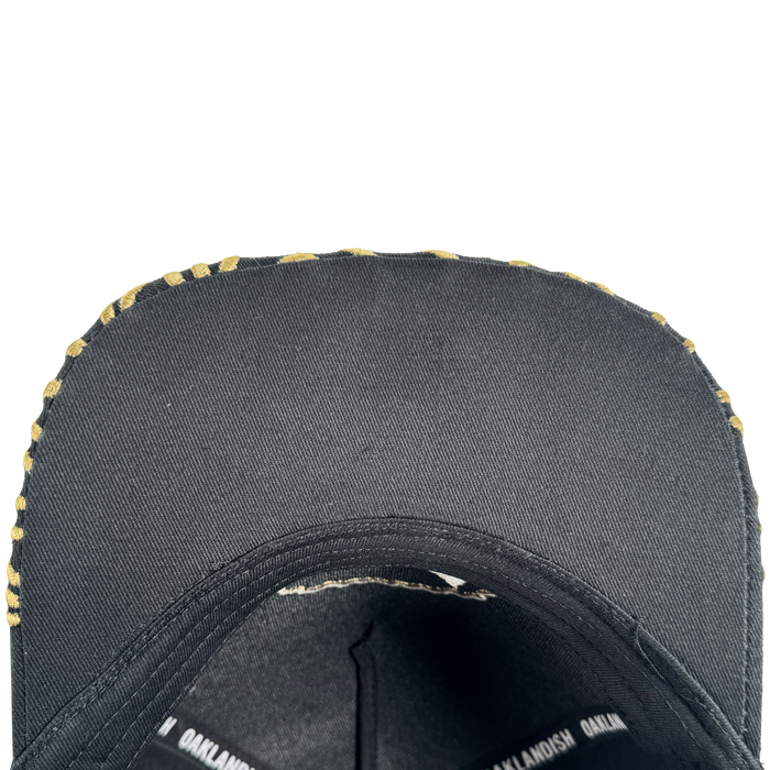 Close-up of the underside of an Oakland Roots SC Growth Cap’s black brim with a yellow camo edge. Inside, black seams and a band with repeating white “OAKLANDISH” text are visible against a white background.