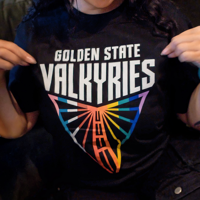 A person points to the GSV Logo Love Tee by GSV, a classic fit, 100% cotton t-shirt with bold “Golden State Valkyries” text and a triangular rainbow wing logo. Only their lower face and hands are shown.