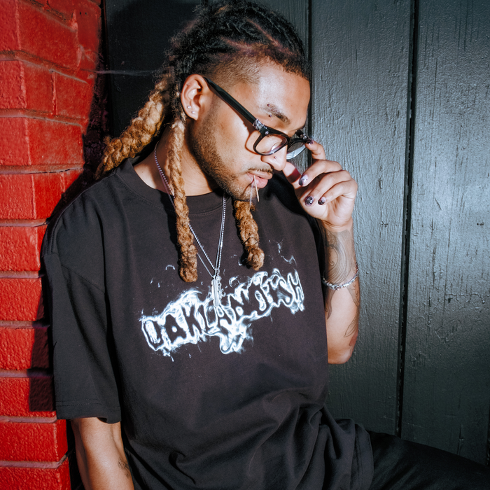 A person with long, braided hair and glasses looks down thoughtfully, touching their glasses. They wear the Oaklandish Haze Tee—a men’s black classic fit t-shirt with bold white text—plus silver necklaces, black nail polish, and stand beside a red brick wall.