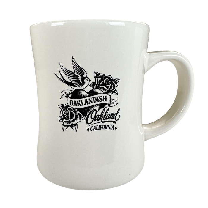 The Heart Diner Mug by Oaklandish is a white 14 oz ceramic mug with black flying bird artwork above an “OAKLANDISH Oakland CALIFORNIA” banner, framed by roses. It features a curved body and large handle for comfortable use.