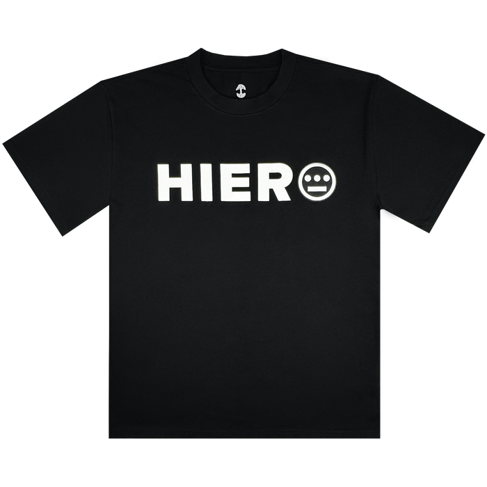 The Hiero Bold Heavy Tee by Hieroglyphics is a black T-shirt with "HIERO" in bold white letters on the chest, inspired by hip-hop culture. It features a face-like white circle with three lines and two dots, round neckline, and short sleeves against a white background.