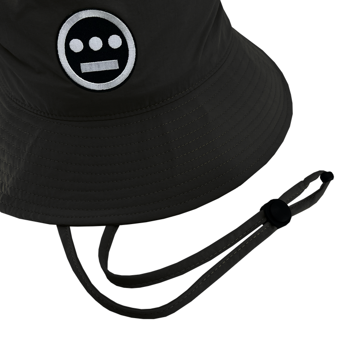 The Hiero Boonie Hat by Hieroglyphics is a dark-colored bucket hat crafted from recycled nylon, featuring a logo with a circular face, two white eyes, and a horizontal bar for a mouth. It includes a chin strap with an adjustable toggle, and the stitching and texture of the fabric are visible.