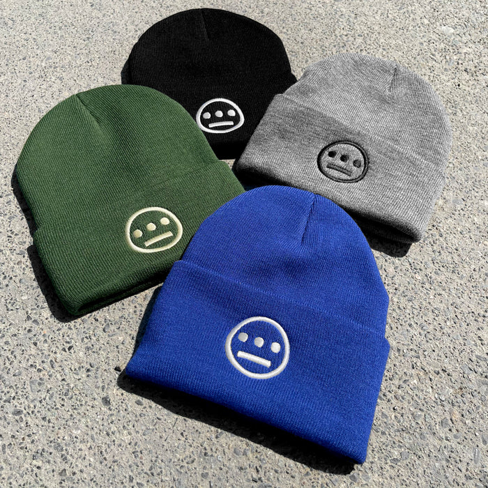 Four Hiero Classic Cuff Beanies by Hieroglyphics are displayed on a concrete surface. This stylish headwear is arranged in a square formation, showcasing colors black, gray, green, and blue. Each beanie features an embroidered circular logo with a three-dot and line design that resembles minimalist hieroglyphics on the front.