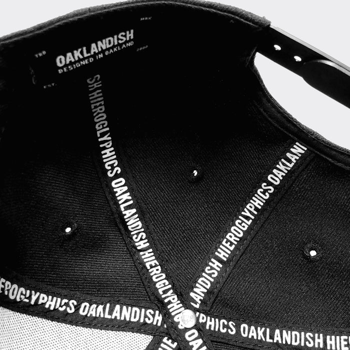 A close-up view of the "Hiero Crew Snapback" cap's interior. The inside labels read "HIERGLYPHICS" repeatedly along the brim and inner seams. "OAKLANDISH DESIGNED IN OAKLAND" is displayed prominently on the inner forehead band. The fabric of this distinctive headwear is attached with metal rivets.
