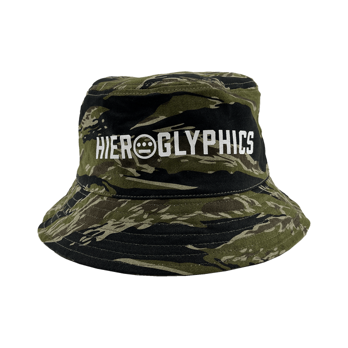The Hieroglyphics Hiero Reversible Camo Bucket is a camouflage bucket hat featuring a green and black tiger camo pattern. The front displays the word "HIEROGLYPHICS" in white, along with a small circular symbol consisting of three dots over a horizontal line within a circle, directly beneath the word.