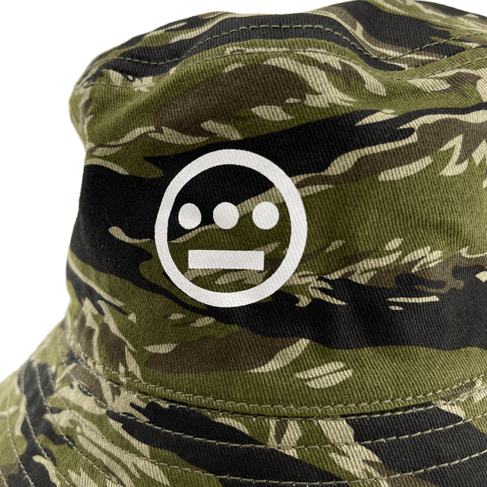 A close-up of the Hieroglyphics Hiero Reversible Camo Bucket hat showcases a white, minimalist face logo. This logo features three filled circles representing eyes and nose, and a horizontal line forming a simplistic mouth. The tiger camouflage pattern incorporates shades of green, brown, and beige.