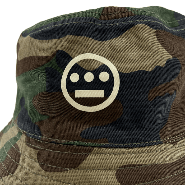Close-up of Hieroglyphics Hip Hop Crew logo on the front crown of a Woodlands Camo bucket hat.